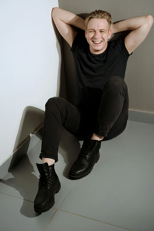 a man sitting on the floor with his hands behind his head, an album cover, by Lucia Peka, pexels contest winner, antipodeans, wearing black boots, smiling slightly, attractive androgynous humanoid, he is wearing a black t-shirt