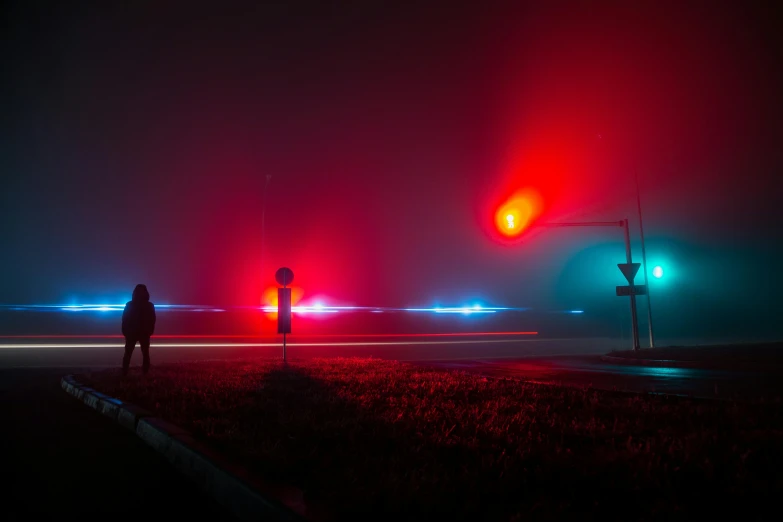 a person standing on the side of a road at night, by Adam Marczyński, red illuminating fog, police lights, foggy volumetric light morning, colorful scene