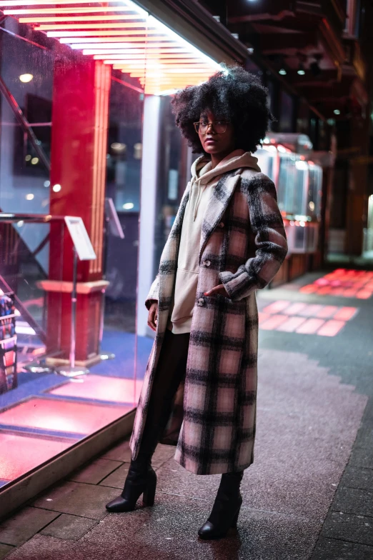 a woman standing in front of a store window, wearing a long coat, tartan hoodie, sza, evening mood