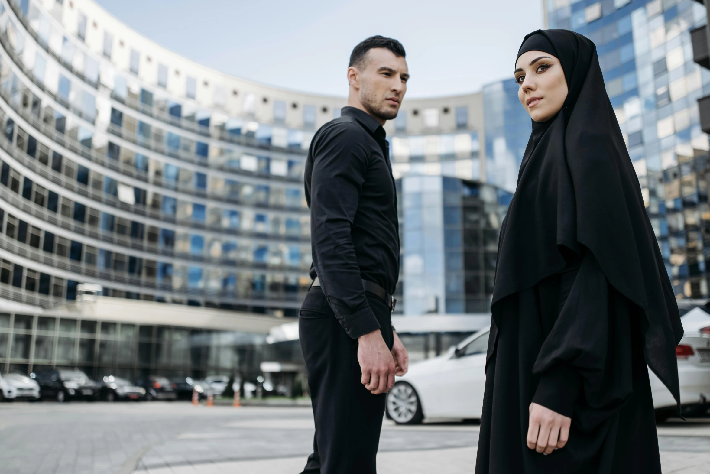 a man and a woman standing in front of a building, by Micha Klein, pexels contest winner, hurufiyya, wearing causal black suits, an arab standing watching over, ( ( theatrical ) ), office clothes