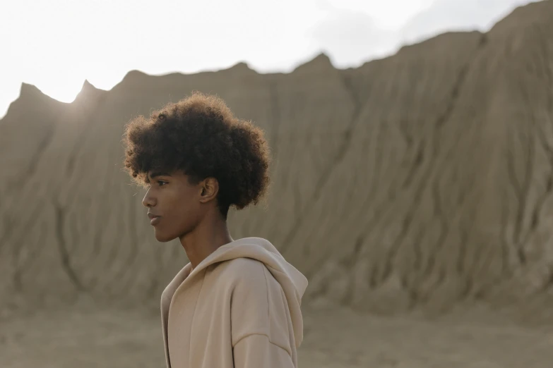 a young man standing in front of a mountain, by Emma Andijewska, trending on pexels, afrofuturism, brown curly hair, black teenage girl, beige hoodie, covered in sand