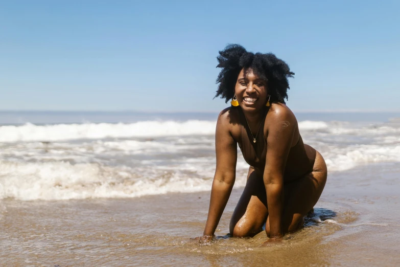 a woman sitting on top of a sandy beach next to the ocean, by Lily Delissa Joseph, pexels contest winner, afrofuturism, standing in a shallow river, cheeks, curls and curves, on a sunny day