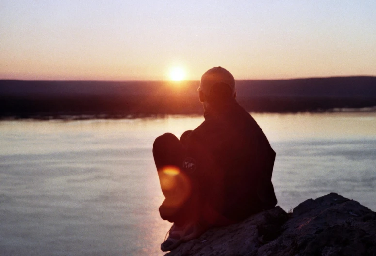a man sitting on top of a rock next to a body of water, by Pamela Ascherson, pexels contest winner, low sun, documentary still, emotional, sittin