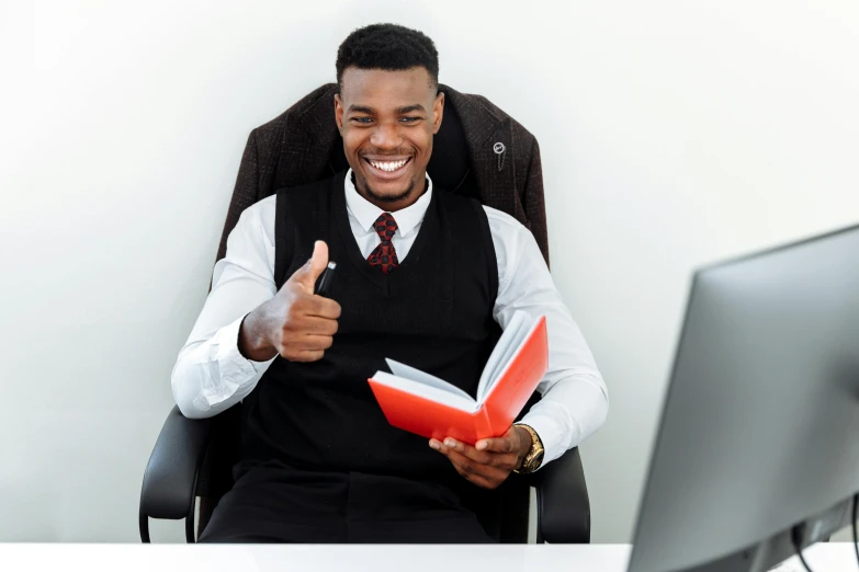 a man sitting in front of a computer giving a thumbs up, pexels contest winner, academic art, black and red suit, black man, holding a book, office furniture