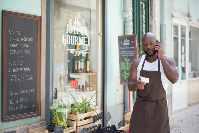 a man standing in front of a store talking on a cell phone, by Barthélemy Menn, pexels contest winner, wearing an apron, kevin garnett, holding an epée, brown