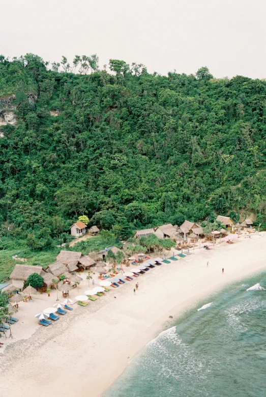 a beach filled with lots of umbrellas next to a lush green hillside, hut, aerial, white sand beach, exterior