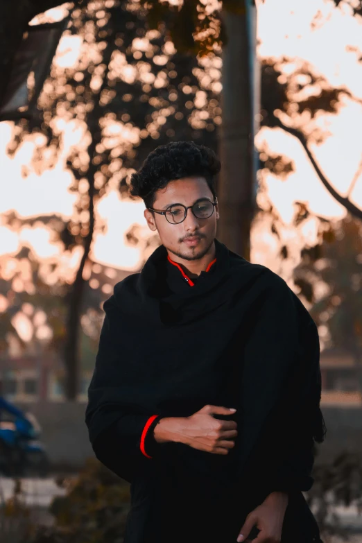 a man with glasses standing in front of a tree, an album cover, inspired by Jitish Kallat, pexels contest winner, realism, wearing black robe, at college, portait photo profile picture, sunset view