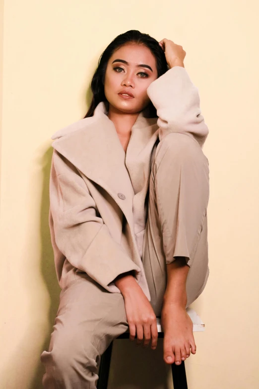 a woman sitting on top of a stool next to a wall, by Robbie Trevino, trending on pexels, dau-al-set, wearing a long beige trench coat, “zendaya, action glamour pose, asian woman