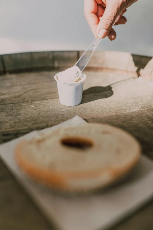 a person dipping something into a cup on top of a wooden barrel, cream, bagels, soft white glow, no crop