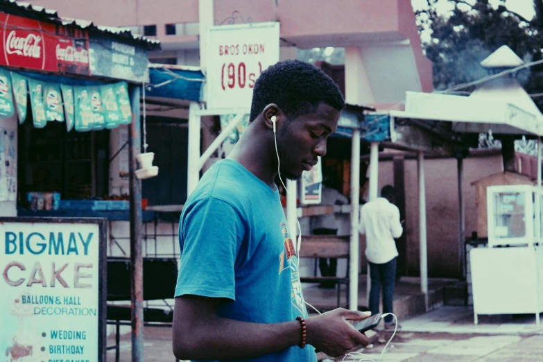 a man in a blue shirt is looking at his cell phone, an album cover, by Dean Ellis, pexels, happening, standing in a township street, with head phones, ( ( dark skin ) ), multi - coloured