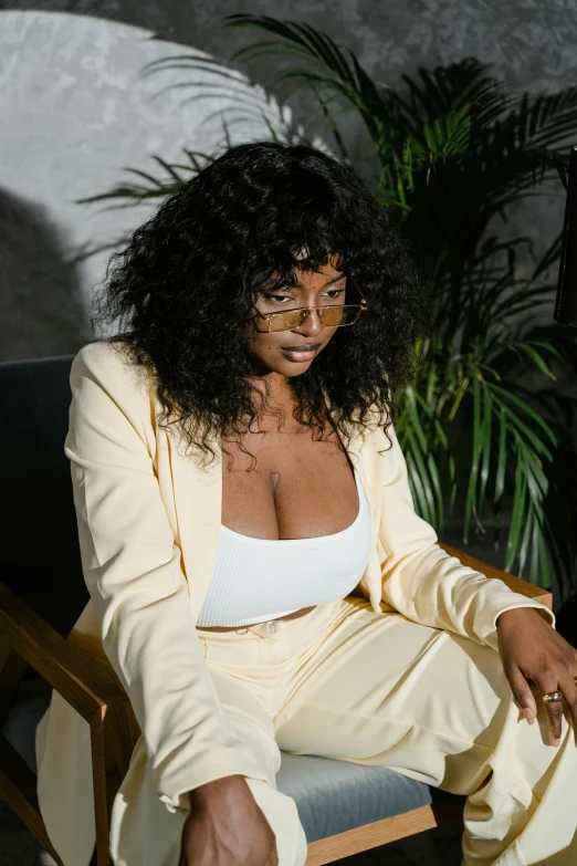 a woman sitting in a chair next to a potted plant, an album cover, inspired by Esaias Boursse, trending on pexels, wearing white suit and glasses, sza, wavy hair yellow theme, seductive stare