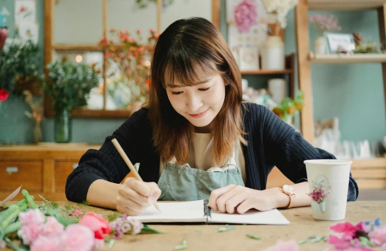 a woman sitting at a table writing on a piece of paper, a drawing, inspired by Kim Jeong-hui, pexels contest winner, japanese flower arrangements, cutest, thumbnail, background image