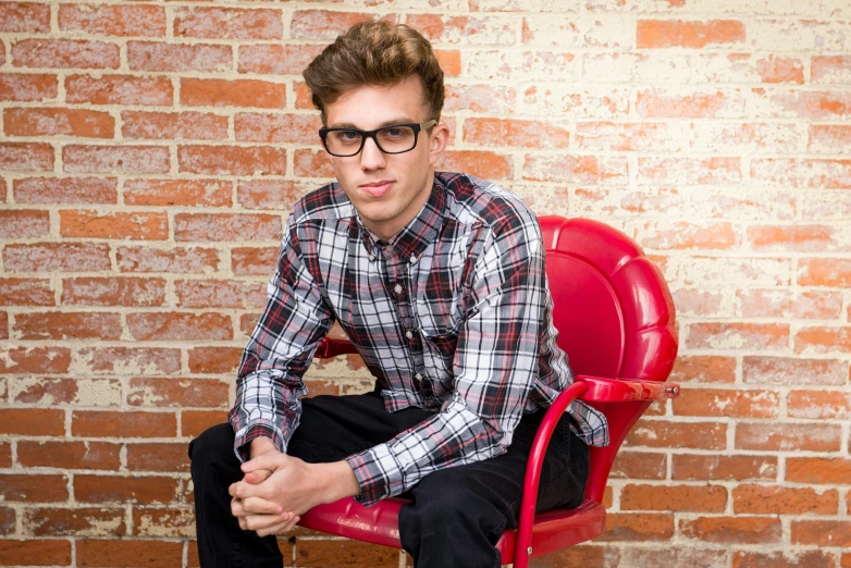 a man sitting on a red chair in front of a brick wall, nerdy, flannel, oliver cook, teenager