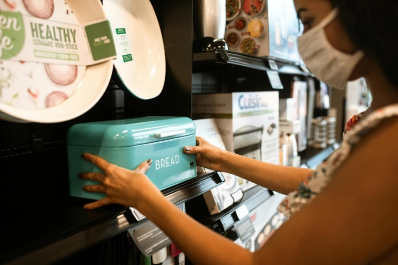 a woman wearing a face mask in a grocery store, by Julia Pishtar, unsplash, happening, white and teal metallic accents, looking at the treasure box, bread, for displaying recipes