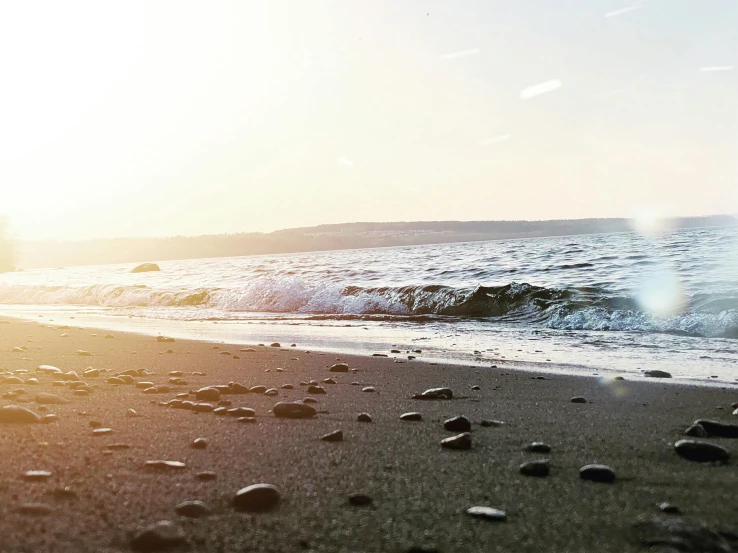 a person riding a surfboard on top of a sandy beach, gravels around, the sea, which shows a beach at sunset, zoomed in