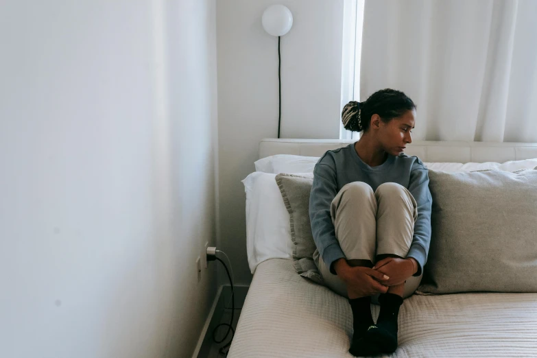 a woman sitting on top of a bed next to a window, trending on pexels, depressed, black teenage girl, grey, tiny person watching