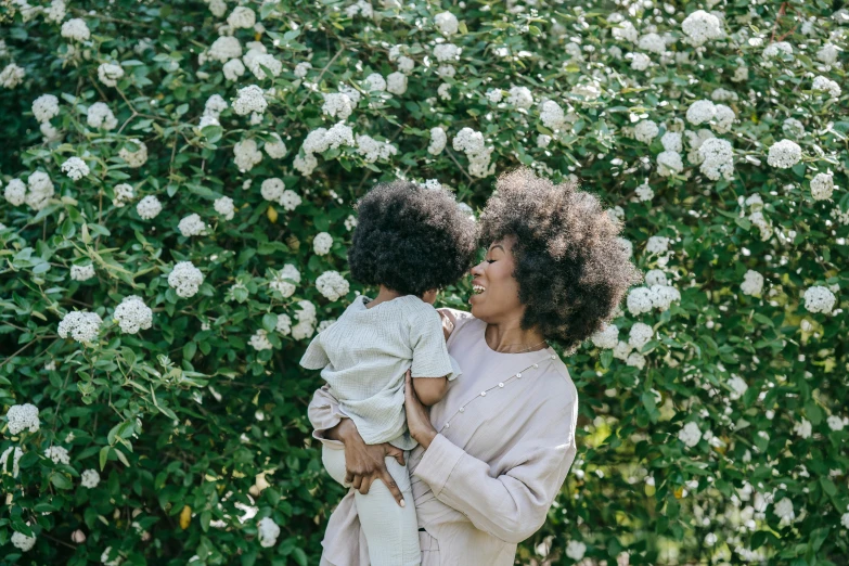 a woman holding a child in front of a bush of flowers, pexels contest winner, curly afro, white hue, lifestyle, [ organic