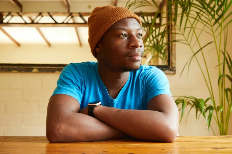 a man sitting at a table with his arms crossed, pexels, wearing teal beanie, very kenyan, avatar image, 2 4 - year - old man