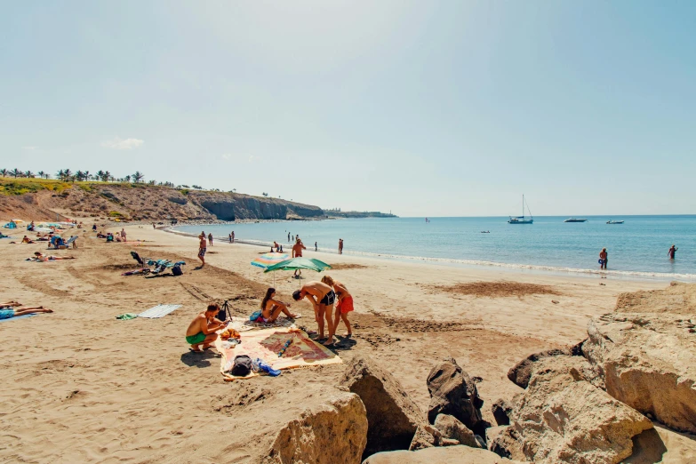 a group of people sitting on top of a sandy beach, near the sea, travel guide, balaskas, sunfaded