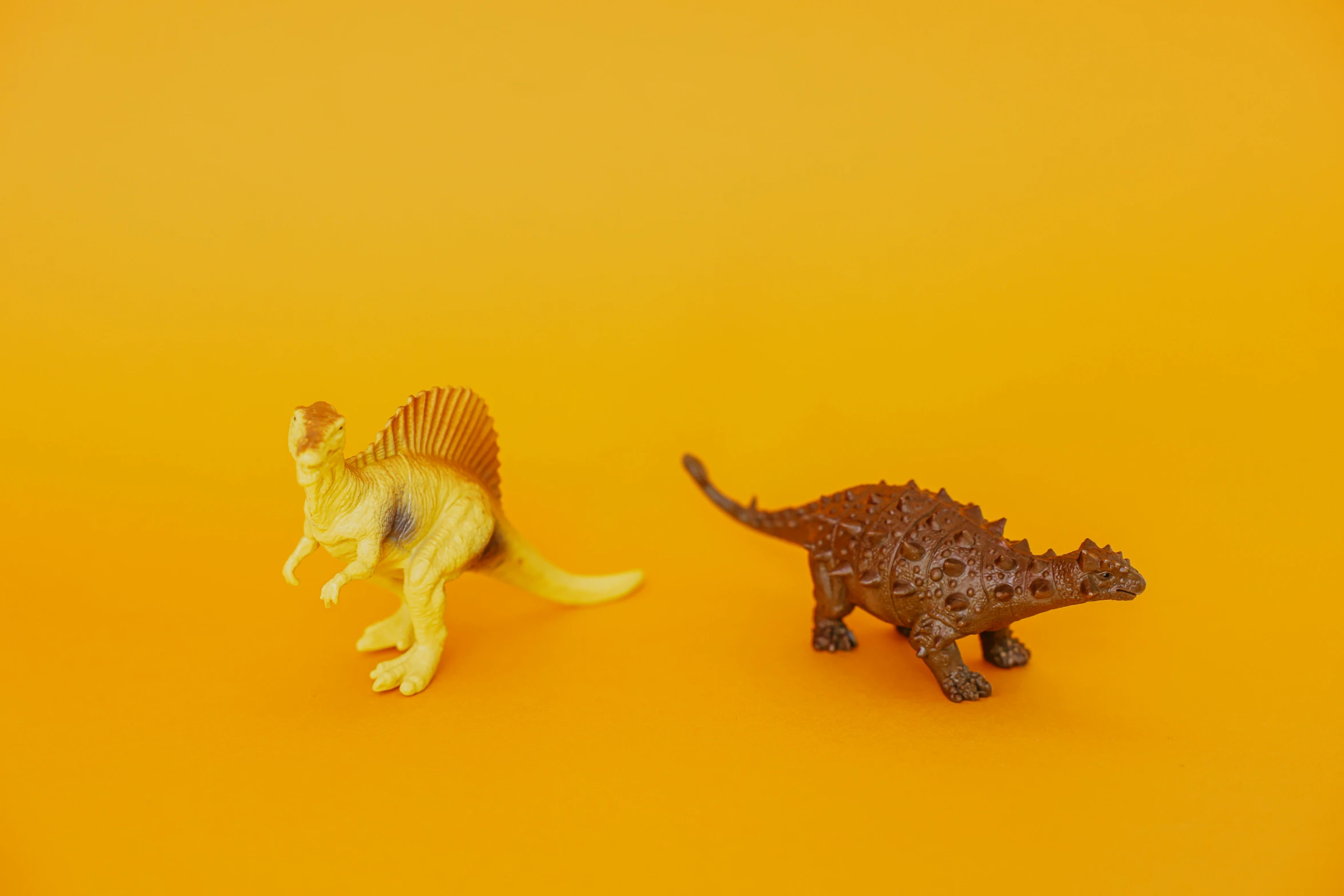 a couple of toy dinosaurs standing next to each other, inspired by Adam Rex, trending on unsplash, yellow backdrop, brown, fossil, 60mm