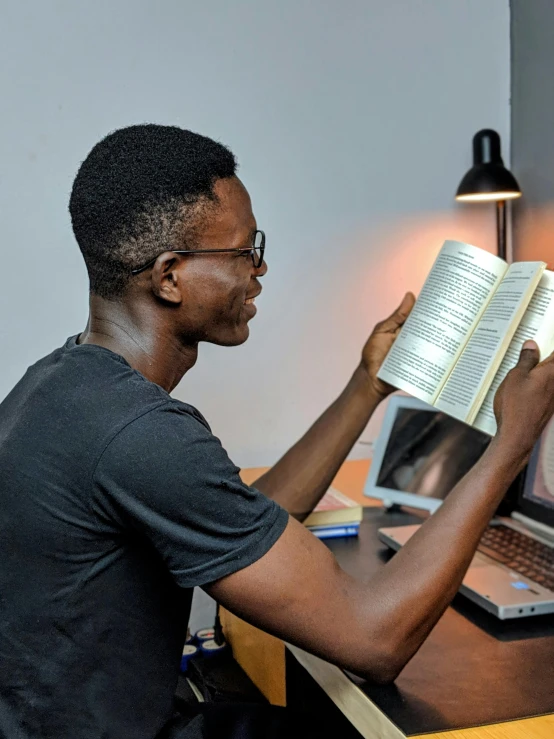 a man sitting at a desk reading a book, pexels contest winner, man is with black skin, profile image, computer science, holding a giant book