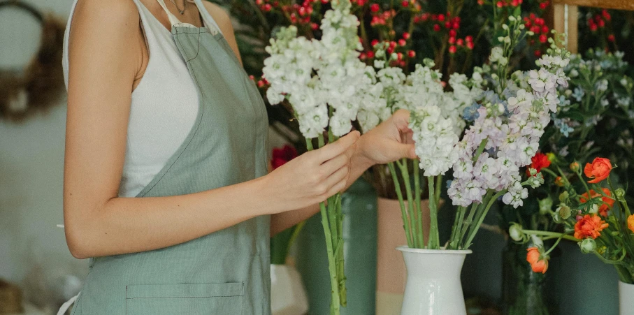 a woman standing in front of a bunch of flowers, trending on pexels, arts and crafts movement, white long gloves, inspect in inventory image, white vase, artist wearing overalls