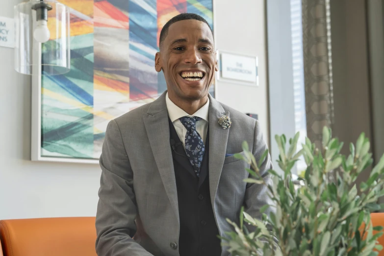 a man sitting on a couch next to a potted plant, a picture, wearing a vest and a tie, brown skin man with a giant grin, professional profile photo, thumbnail