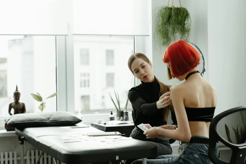 two women sitting at a table in front of a window, a tattoo, trending on pexels, with a stethoscope, black and red hair, showing her shoulder from back, colourised