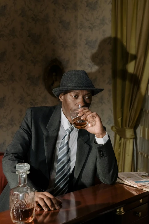 a man sitting at a desk drinking from a glass, an album cover, inspired by Gordon Parks, lance reddick, [ theatrical ], movie still 8 k, fedora