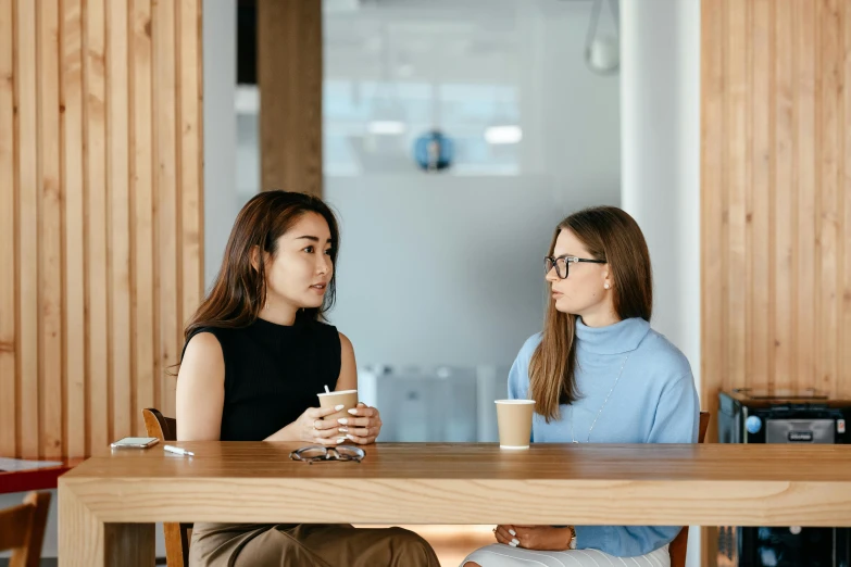 two women sitting at a table talking to each other, trending on pexels, aussie baristas, sitting on top a table, brown, without duplicate image