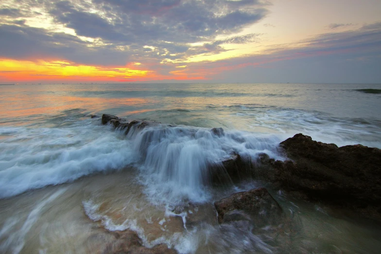 a large body of water sitting on top of a sandy beach, a picture, during a sunset, splashing water, rocky shore, slide show