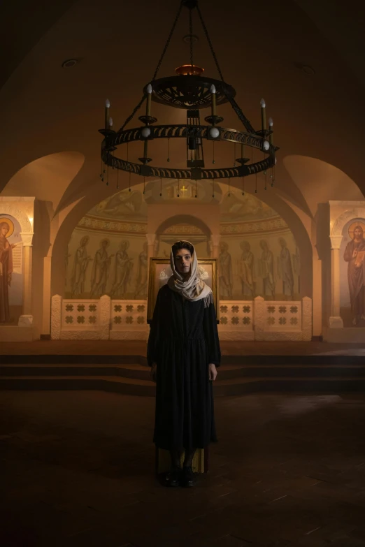 a woman standing in front of a chandelier in a church, inspired by Andrei Rublev, real life photo of a syrian man, national geographic channel, robed, [ theatrical ]