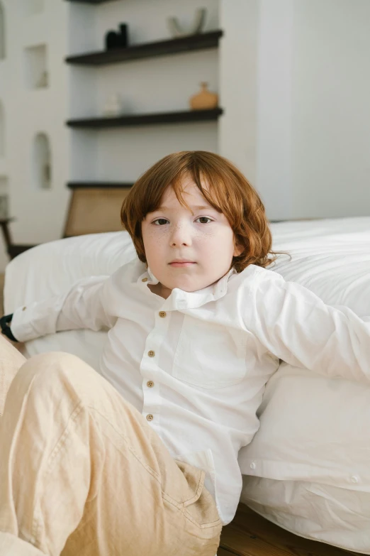 a little boy that is laying on a bed, wearing a white shirt, sitting on a couch, looking confident, concerned