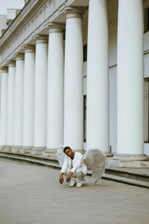 a man kneeling on the ground in front of a building, by Cosmo Alexander, pexels contest winner, visual art, large white wings, colonnade, doing a sassy pose, young wan angel