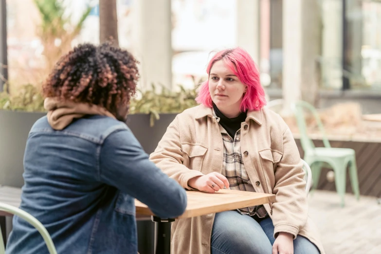 two women sitting at a table talking to each other, by Nick Fudge, trending on pexels, with pink hair, casually dressed, in an urban setting, warm coloured