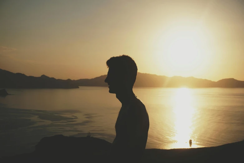 a man standing next to a body of water at sunset, pexels contest winner, profile picture 1024px, sunfaded, outlined silhouettes, sitting