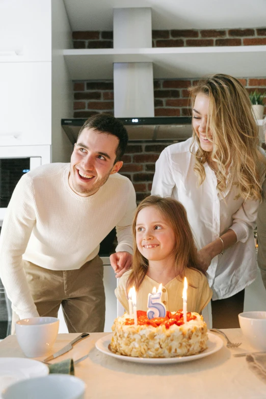 a group of people standing around a table with a cake, with a kid, white candles, profile image, happy lighting