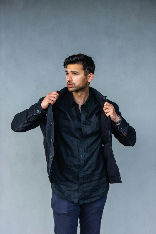 a man standing in front of a gray wall, rugged black clothes, wearing hunter coat, nathan fielder, all black matte product