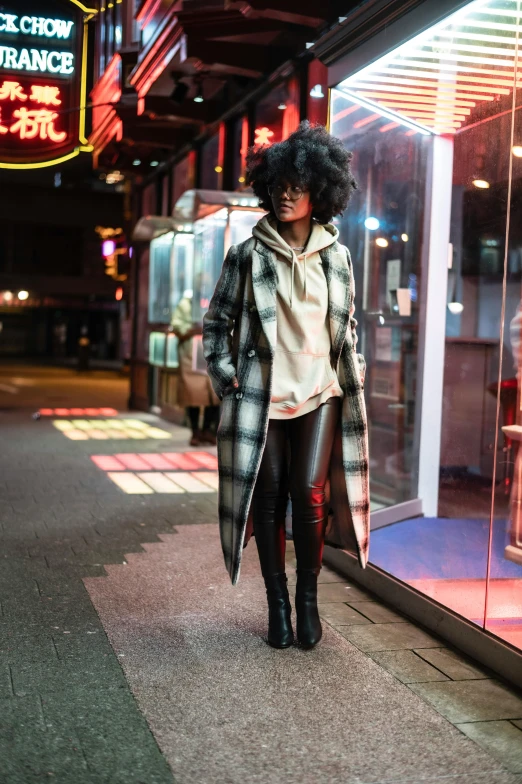 a woman standing on a sidewalk in front of a store, stylish coat for a rave, flannel, night life, black young woman