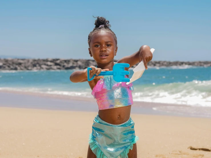 a little girl standing on top of a sandy beach, with blue skin, orisha, wave a hand at the camera, wearing pearl neon bikini