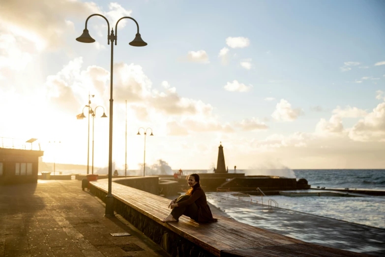 a woman sitting on a bench next to the ocean, by Niko Henrichon, pexels contest winner, happening, cinematic lens flare, azores, city morning, girl under lantern
