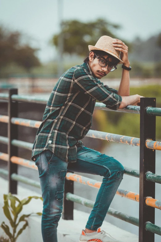 a man in a hat leaning on a railing, a picture, inspired by Rudy Siswanto, trending on pexels, sumatraism, wearing plaid shirt, zayn malik, confident relaxed pose, green and brown clothes