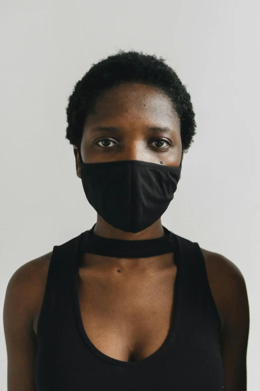 a woman wearing a black face mask, by Nina Hamnett, she is wearing a black tank top, dark complexion, on grey background, environment