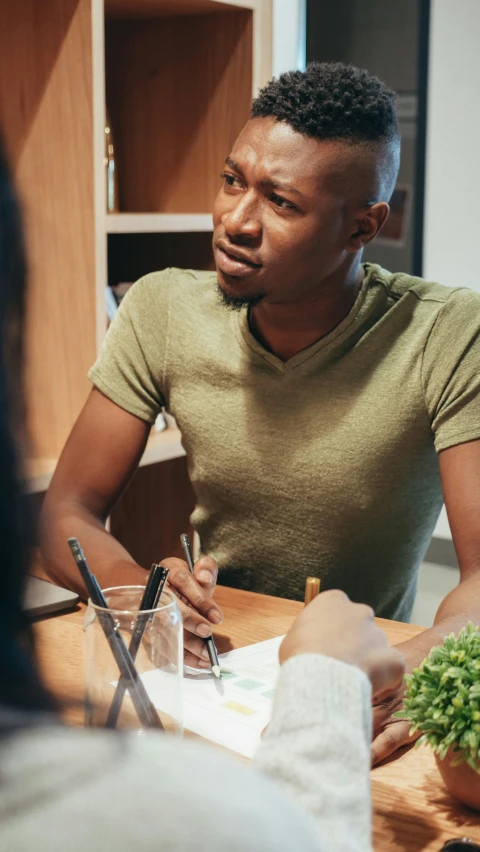 a group of people sitting around a wooden table, trending on pexels, african man, pondering, in a dark green polo shirt, lgbtq