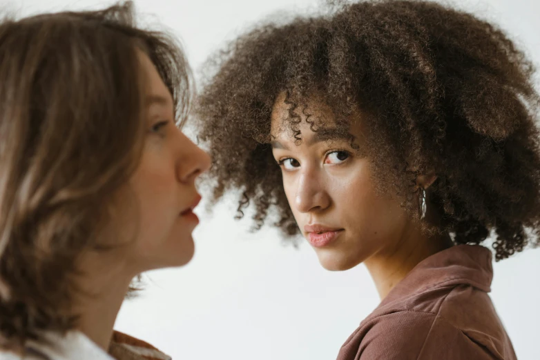 two women standing next to each other looking at each other, trending on pexels, brown curly hair, pouty, black young woman, realistic »