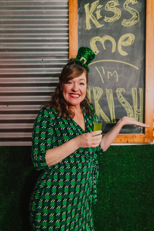 a woman in a green dress standing in front of a chalkboard, holding a drink, irish genes, wearing green suit, fan favorite