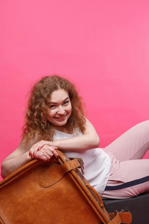 a woman sitting on top of a piece of luggage, brown curly hair, ((pink)), holding a leather purse, smiling coyly