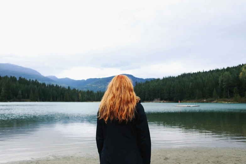 a woman standing on a beach next to a body of water, pexels contest winner, long strawberry - blond hair, mountain lakes, slight overcast weather, view from behind