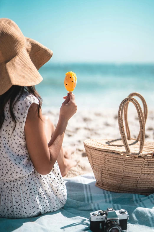 a woman sitting on top of a beach next to a basket, eating ice cream, yellow parasol, clear and sunny, holding maracas