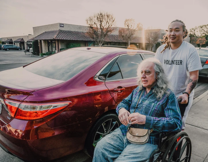 a man sitting in a wheel chair next to a woman, a portrait, by Michael Goldberg, pexels contest winner, happening, standing in front of lowrider, carpool karaoke, avatar image, asian male